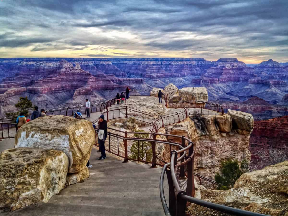 A paved South Rim path winds between stone railings and visitors in Grand Canyon National Park, Arizona, United States.