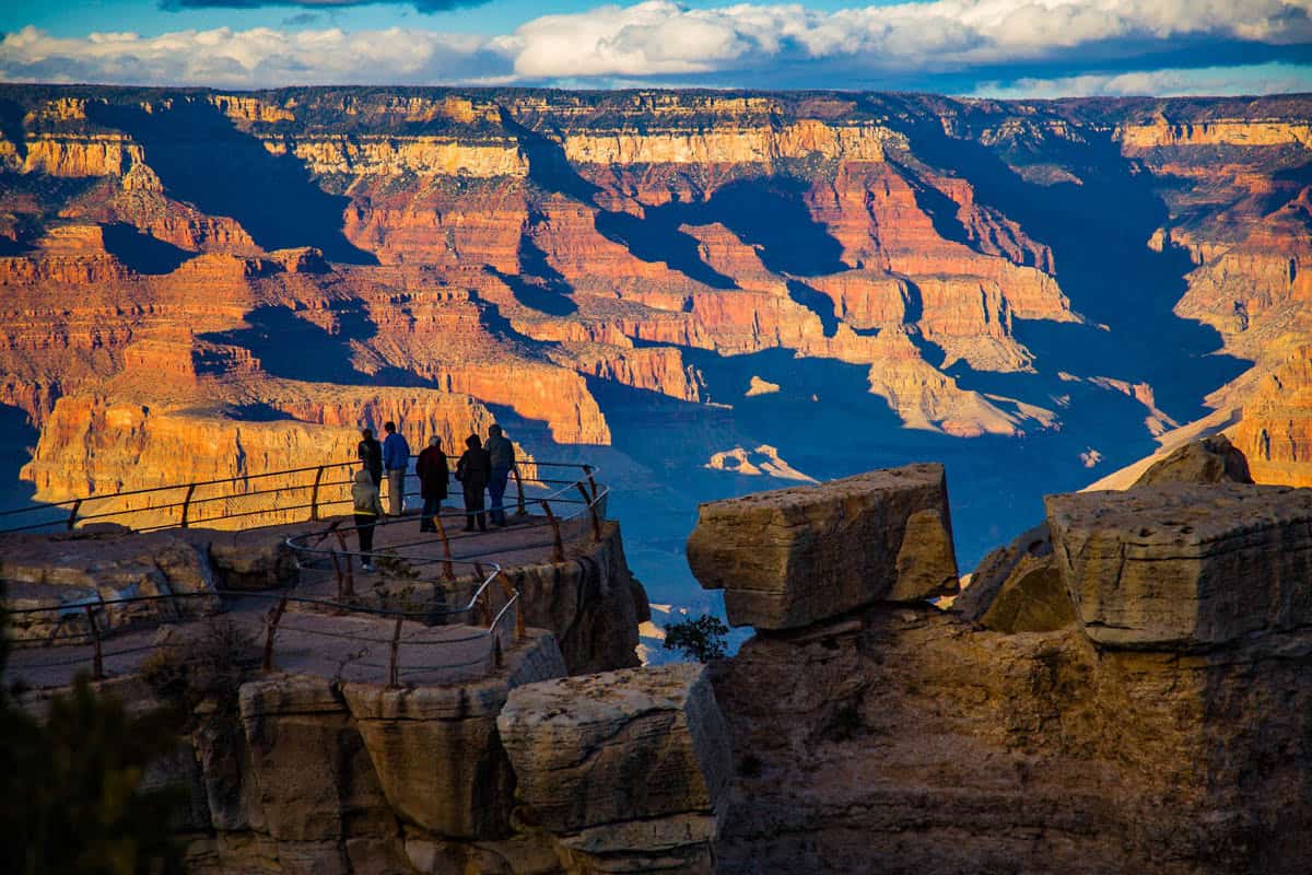 Visitors standing along a rocky South Rim overlook in Grand Canyon National Park, with Sunlit canyon walls visible.